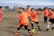 A group of young children playing soccer on a grassy field. They are wearing bright orange jerseys with black shorts, focusing intently on the game as they chase a soccer ball. The sky is partly cloudy, and there are trees and other players visible in the background.