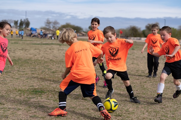 A group of young children playing soccer on a grassy field. They are wearing bright orange jerseys with black shorts, focusing intently on the game as they chase a soccer ball. The sky is partly cloudy, and there are trees and other players visible in the background.