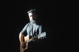 A person with a beard plays an acoustic guitar under a spotlight, surrounded by darkness. The focus is on the musician, creating a dramatic and intimate atmosphere.