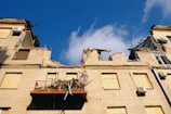 A damaged building with severe structural damage is apparent, showcasing crumbling walls and destroyed sections. The windows are boarded up, and debris is scattered on a collapsed balcony. The sky is clear and blue.