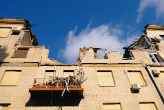 A damaged building with severe structural damage is apparent, showcasing crumbling walls and destroyed sections. The windows are boarded up, and debris is scattered on a collapsed balcony. The sky is clear and blue.