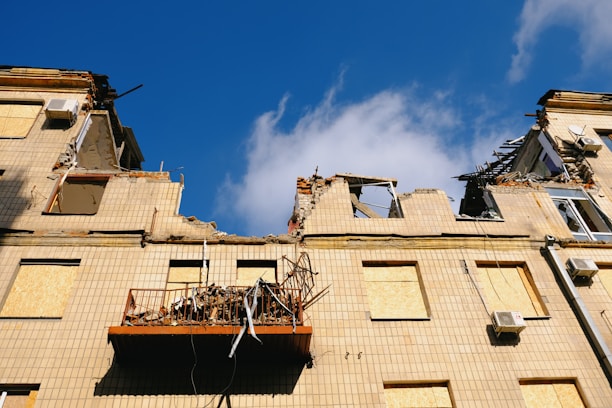 A damaged building with severe structural damage is apparent, showcasing crumbling walls and destroyed sections. The windows are boarded up, and debris is scattered on a collapsed balcony. The sky is clear and blue.