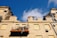 A damaged building with severe structural damage is apparent, showcasing crumbling walls and destroyed sections. The windows are boarded up, and debris is scattered on a collapsed balcony. The sky is clear and blue.
