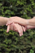 Several hands gently resting on top of one another against a blurred natural green background. The hands convey a sense of unity and support.
