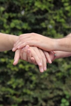 Several hands gently resting on top of one another against a blurred natural green background. The hands convey a sense of unity and support.