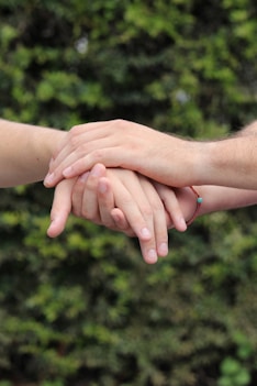 Several hands gently resting on top of one another against a blurred natural green background. The hands convey a sense of unity and support.