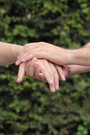 Several hands gently resting on top of one another against a blurred natural green background. The hands convey a sense of unity and support.
