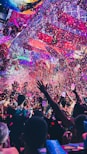 A vibrant indoor event with colorful confetti filling the air. A large crowd of people are gathered, many with raised arms and smartphones capturing the moment. The atmosphere is lively with dynamic lighting casting hues of purple, pink, and blue over the scene.