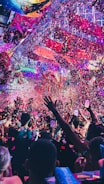 A vibrant indoor event with colorful confetti filling the air. A large crowd of people are gathered, many with raised arms and smartphones capturing the moment. The atmosphere is lively with dynamic lighting casting hues of purple, pink, and blue over the scene.