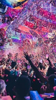 A vibrant indoor event with colorful confetti filling the air. A large crowd of people are gathered, many with raised arms and smartphones capturing the moment. The atmosphere is lively with dynamic lighting casting hues of purple, pink, and blue over the scene.