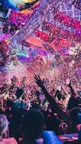 A vibrant indoor event with colorful confetti filling the air. A large crowd of people are gathered, many with raised arms and smartphones capturing the moment. The atmosphere is lively with dynamic lighting casting hues of purple, pink, and blue over the scene.