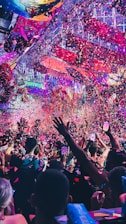 A vibrant indoor event with colorful confetti filling the air. A large crowd of people are gathered, many with raised arms and smartphones capturing the moment. The atmosphere is lively with dynamic lighting casting hues of purple, pink, and blue over the scene.