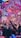 A vibrant indoor event with colorful confetti filling the air. A large crowd of people are gathered, many with raised arms and smartphones capturing the moment. The atmosphere is lively with dynamic lighting casting hues of purple, pink, and blue over the scene.