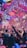 A vibrant indoor event with colorful confetti filling the air. A large crowd of people are gathered, many with raised arms and smartphones capturing the moment. The atmosphere is lively with dynamic lighting casting hues of purple, pink, and blue over the scene.