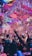 A vibrant indoor event with colorful confetti filling the air. A large crowd of people are gathered, many with raised arms and smartphones capturing the moment. The atmosphere is lively with dynamic lighting casting hues of purple, pink, and blue over the scene.