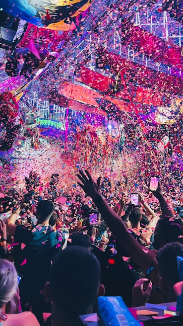 A vibrant indoor event with colorful confetti filling the air. A large crowd of people are gathered, many with raised arms and smartphones capturing the moment. The atmosphere is lively with dynamic lighting casting hues of purple, pink, and blue over the scene.