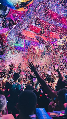 A vibrant indoor event with colorful confetti filling the air. A large crowd of people are gathered, many with raised arms and smartphones capturing the moment. The atmosphere is lively with dynamic lighting casting hues of purple, pink, and blue over the scene.