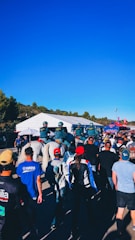 A group of people walking towards a large white tent, with several police officers on horseback guiding the crowd. Many individuals are wearing caps and jackets, with some branded apparel visible. The sky is clear and blue, and the surrounding area includes greenery and some structures.