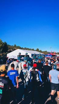 A group of people walking towards a large white tent, with several police officers on horseback guiding the crowd. Many individuals are wearing caps and jackets, with some branded apparel visible. The sky is clear and blue, and the surrounding area includes greenery and some structures.