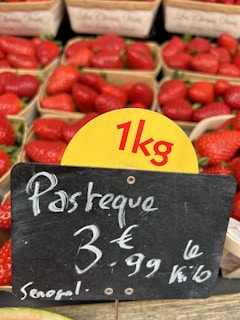 Baskets of strawberries are displayed in a market setting with a sign indicating a price of 3.99 euros per kilogram, with the product labeled as originating from Senegal. A small portion of what appears to be watermelon is visible at the bottom of the image.