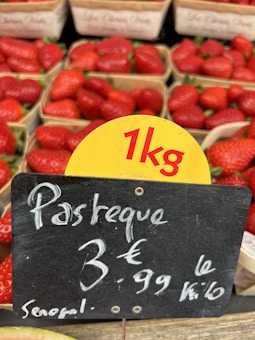 Baskets of strawberries are displayed in a market setting with a sign indicating a price of 3.99 euros per kilogram, with the product labeled as originating from Senegal. A small portion of what appears to be watermelon is visible at the bottom of the image.