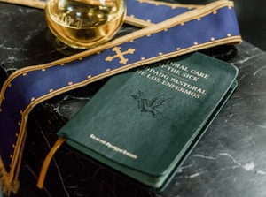 A dark marble table holds a book titled 'Pastoral Care of the Sick' with bilingual text. A purple stole with gold embroidery and a small glass container filled with liquid are placed nearby.