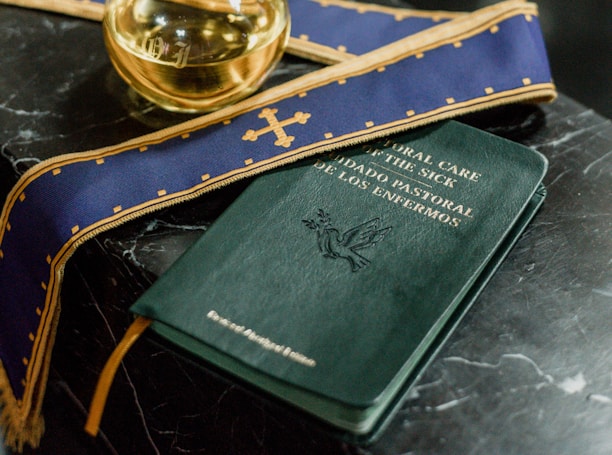 A dark marble table holds a book titled 'Pastoral Care of the Sick' with bilingual text. A purple stole with gold embroidery and a small glass container filled with liquid are placed nearby.