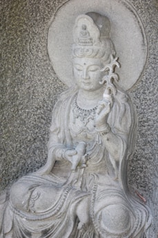 A stone statue of a serene deity or bodhisattva with intricate detailing, seated with one hand holding a scroll and the other raised, holding a twig-like object. The figure has a peaceful expression, adorned with bead necklaces and a headdress featuring a smaller carved figure.