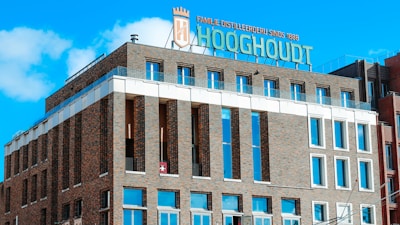 A brown brick building with multiple rows of rectangular windows and a rooftop sign displaying 'Hooghoudt' along with other text. The architecture has a modern design, featuring white accents and glass surfaces. The sky is clear and blue with a few clouds.