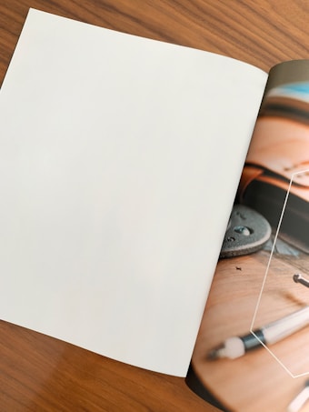 A magazine is open on a wooden surface. The left page is blank, while the right page shows a close-up of a skateboard with a metal part and wheels, and part of a pen is visible. The image is partially blurred, giving it an artistic touch.