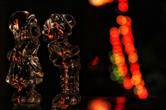 Two glass figurines in a pose that suggests affection are illuminated by warm lights, creating an intimate and cozy atmosphere. In the background, bokeh circles from colorful lights blur into red, orange, and green, adding depth and festive ambiance.