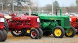 A row of vintage tractors in various colors is parked on a gravel and grass surface near a chain-link fence. The foreground features a red Farmall tractor and a green John Deere tractor with bright yellow wheels. Additional tractors in red can be seen in the background. The setting appears to be an outdoor event, as indicated by the small signs attached to the tractors and a few people interacting with them.