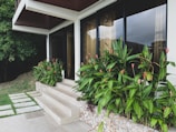 A contemporary house exterior features large glass windows with sheer curtains, providing a view of the interior. The entrance area includes a stone staircase leading up to the door. Lush green tropical plants with vibrant red flowers line the building, contrasting with the beige stone tiles. A well-manicured lawn and a dense tree are visible on the left side.