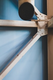 A section of a metal structure with riveted joints and braces against a light blue background. The metal parts appear to be aged with some rust and grime, particularly around the joints and edges.