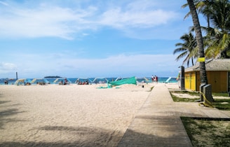 A sandy beach with several reclining lounge chairs under blue canopies. People are relaxing on the beach, and palm trees provide a tropical atmosphere. A yellow building with a tiled roof is located to the right next to a boardwalk. The ocean is visible in the background under a partly cloudy blue sky.