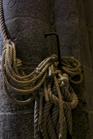 Thick ropes are coiled and hanging on a sturdy metal hook embedded into a textured stone wall. The ropes appear aged, with frayed ends, suggesting heavy use or age. The scene is dimly lit, casting subtle shadows on the ropes and the wall, emphasizing the rugged texture of both materials.
