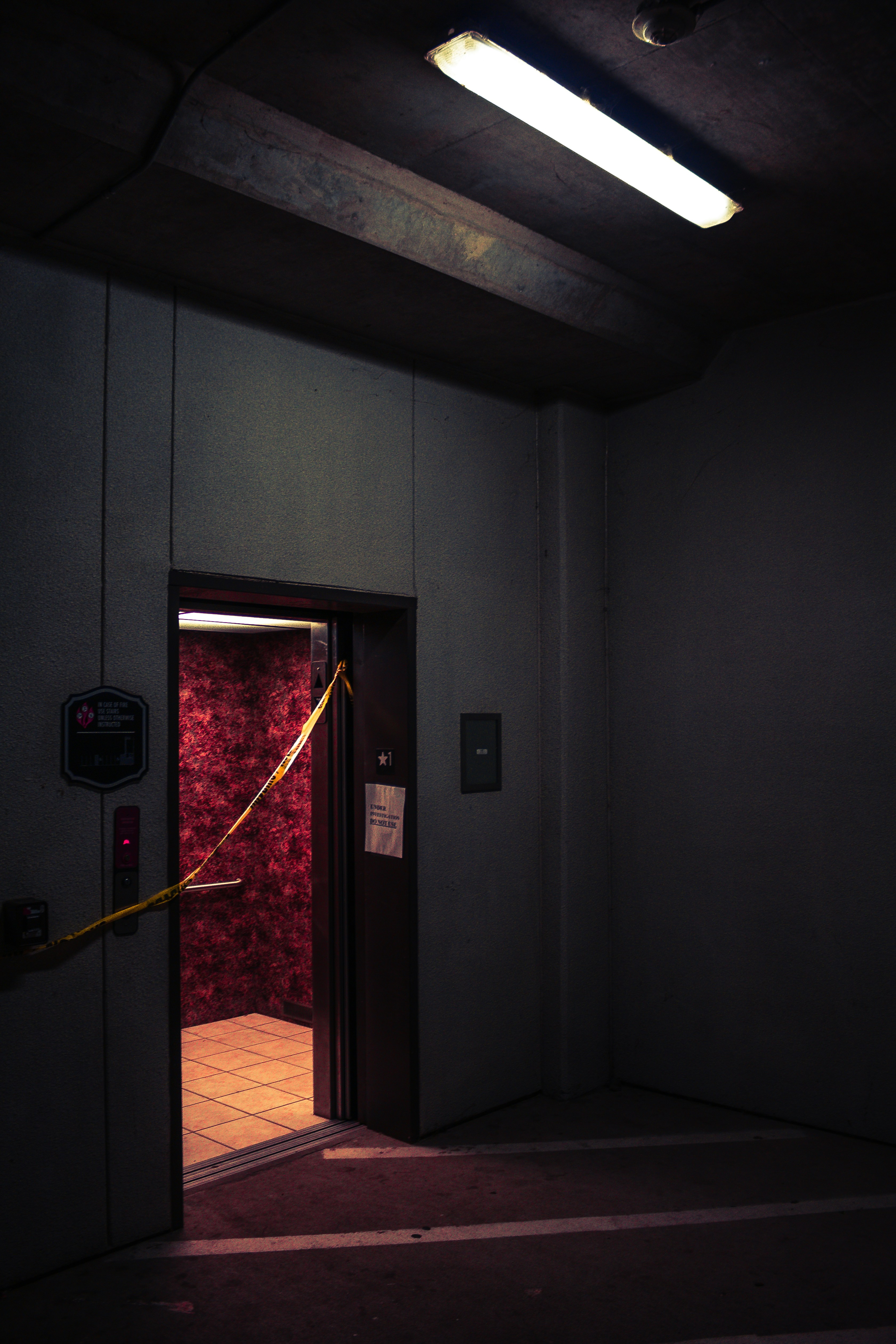 A dimly lit elevator with an open door in a parking garage or industrial space. The walls inside the elevator have a textured red pattern, and there is yellow caution tape stretched across the doorway. Overhead, a fluorescent light casts a harsh glow, contributing to the gloomy atmosphere.