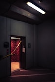A dimly lit elevator with an open door in a parking garage or industrial space. The walls inside the elevator have a textured red pattern, and there is yellow caution tape stretched across the doorway. Overhead, a fluorescent light casts a harsh glow, contributing to the gloomy atmosphere.