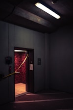 A dimly lit elevator with an open door in a parking garage or industrial space. The walls inside the elevator have a textured red pattern, and there is yellow caution tape stretched across the doorway. Overhead, a fluorescent light casts a harsh glow, contributing to the gloomy atmosphere.