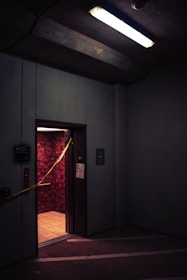 A dimly lit elevator with an open door in a parking garage or industrial space. The walls inside the elevator have a textured red pattern, and there is yellow caution tape stretched across the doorway. Overhead, a fluorescent light casts a harsh glow, contributing to the gloomy atmosphere.