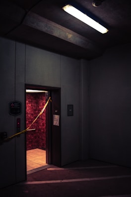A dimly lit elevator with an open door in a parking garage or industrial space. The walls inside the elevator have a textured red pattern, and there is yellow caution tape stretched across the doorway. Overhead, a fluorescent light casts a harsh glow, contributing to the gloomy atmosphere.