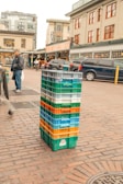 A stack of multi-colored plastic crates is placed on a cobblestone street in front of a bustling marketplace. In the background, a market sign is visible above a row of parked cars, and pedestrians are walking nearby. A person wearing a dark jacket and cap stands to the left. The area has an urban and lively atmosphere.