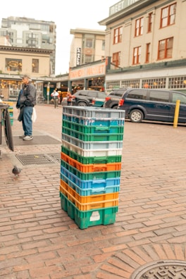 A stack of multi-colored plastic crates is placed on a cobblestone street in front of a bustling marketplace. In the background, a market sign is visible above a row of parked cars, and pedestrians are walking nearby. A person wearing a dark jacket and cap stands to the left. The area has an urban and lively atmosphere.
