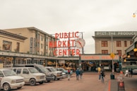 A busy urban street scene outside a market. The large, red neon sign reads 'Public Market Center' with a classic clock next to it. Several vehicles are parked in front of the market entrance. People are walking towards the entrance, where another sign says 'Farmers Market.' Buildings surround the market, including a hotel on the left.
