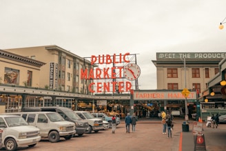 A busy urban street scene outside a market. The large, red neon sign reads 'Public Market Center' with a classic clock next to it. Several vehicles are parked in front of the market entrance. People are walking towards the entrance, where another sign says 'Farmers Market.' Buildings surround the market, including a hotel on the left.