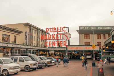 A busy urban street scene outside a market. The large, red neon sign reads 'Public Market Center' with a classic clock next to it. Several vehicles are parked in front of the market entrance. People are walking towards the entrance, where another sign says 'Farmers Market.' Buildings surround the market, including a hotel on the left.