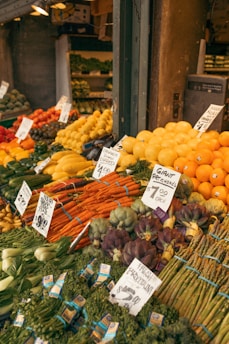 A vibrant market stall displays a wide array of fresh fruits and vegetables, including oranges, bananas, artichokes, carrots, and asparagus. Each produce has a handwritten label with prices attached, enhancing the authenticity and charm of the market setting.