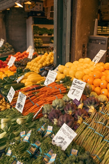 A vibrant market stall displays a wide array of fresh fruits and vegetables, including oranges, bananas, artichokes, carrots, and asparagus. Each produce has a handwritten label with prices attached, enhancing the authenticity and charm of the market setting.
