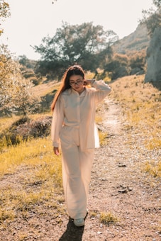 A person with long hair and glasses is walking along a sunlit path in a natural setting. They are wearing a light-colored outfit and appear relaxed, with one hand in their hair. The background features trees, hills, and a few rocks, with sunlight casting warm tones over the scene.