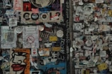 A dense collage of various stickers and graffiti covers a wall. The wall features a mix of colorful and monochrome stickers, tags, and small posters overlapping each other. Among the visual elements are bold text, stylized designs, and hand-drawn elements. Some stickers contain messages, logos, and artistic illustrations.